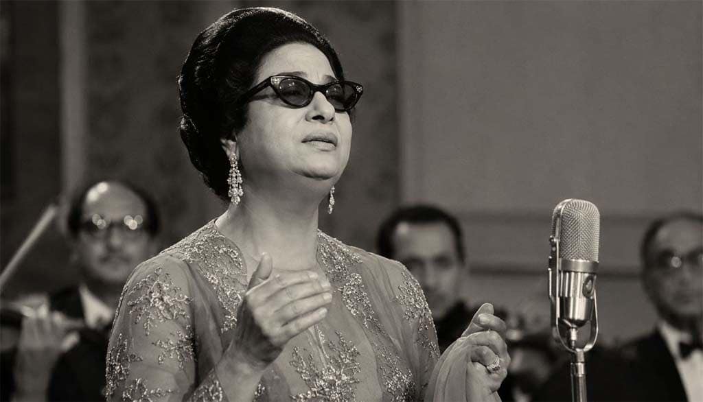 صورة واقعية بالأبيض والأسود للفنانة أم كلثوم أثناء الغناء على المسرح وسط فرقة موسيقية بطابع كلاسيكي قديم.